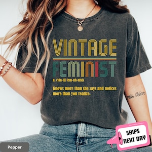 May include: Dark gray t-shirt with the words "VINTAGE FEMINIST" in a retro font. The shirt includes a definition and a quote about feminism. The shirt is a comfortable, casual style.