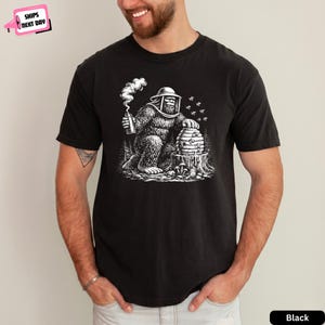 May include: A black t-shirt featuring a white graphic illustration of a Bigfoot creature wearing a beekeeper hat and veil. The creature is holding a smoker and tending to a beehive resting on a tree stump, surrounded by flying bees and forest trees.