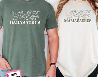 Könnte beinhalten: Ein moosgrünes T-Shirt mit weißer Strichzeichnung von Dinosauriern und dem Text DADASAURUS sowie ein elfenbeinfarbenes T-Shirt mit dunkelgrauer Strichzeichnung von Dinosauriern und dem Text MAMASAURUS. Beide T-Shirts haben kurze Ärmel und einen Rundhalsausschnitt.