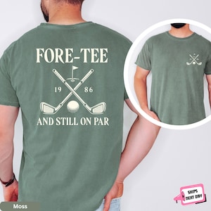 May include: A moss green t-shirt featuring a large back graphic with two crossed golf clubs, a golf ball, a flag, and the text "FORE-TEE 1986 AND STILL ON PAR". The front chest area displays a smaller version of the crossed golf clubs graphic.