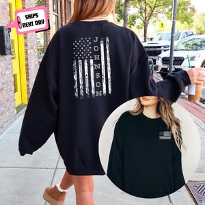 May include: Black sweatshirt featuring a distressed American flag design with the name "JOHNSON" vertically printed on the right side. The design is in white. A smaller version of the flag is on the front of the sweatshirt. The sweatshirt is a casual apparel item.