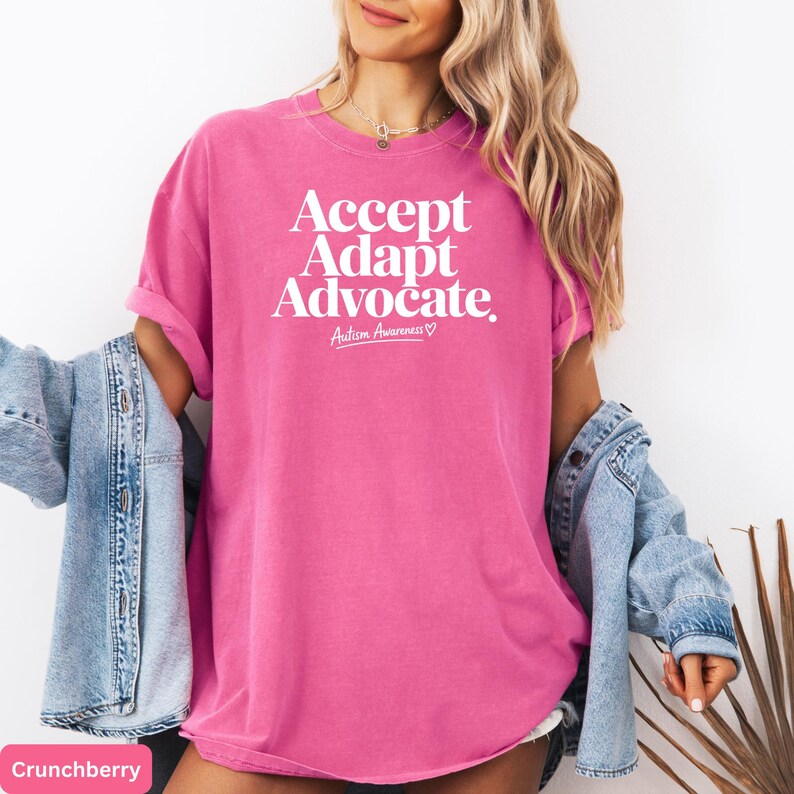 A woman wearing a pink t-shirt with the text "Accept Adapt Advocate" printed on it, standing against a white background.