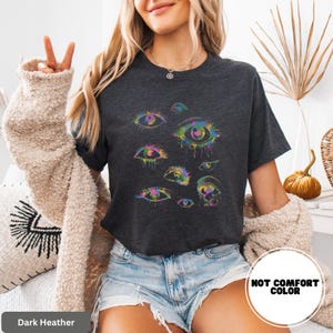 A woman wearing a black t-shirt with a colorful graphic of eyes and a peace sign, paired with denim shorts and a beige cardigan.