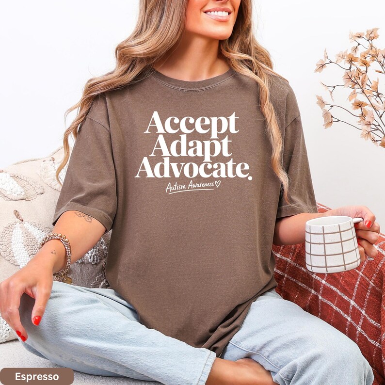 A woman wearing a brown t-shirt with the text "Accept Adapt Advocate Autism Awareness" is sitting on a couch, holding a coffee mug.