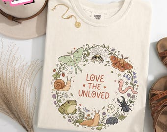 Può includere: Una t-shirt color avorio con un'illustrazione circolare di vari insetti e piccoli animali, tra cui una falena, un ragno, una lumaca, una rana e uno scarabeo, che circondano il testo "LOVE THE UNLOVED" in un carattere marrone arrotondato.
