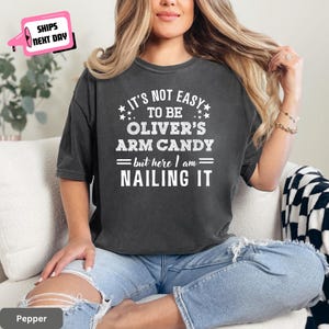 May include: Pepper-colored t-shirt with the text "IT'S NOT EASY TO BE OLIVER'S ARM CANDY = but here I am = NAILING IT" in white. The shirt is worn with blue jeans. A pink and white "SHIPS NEXT DAY" graphic is in the upper left corner.
