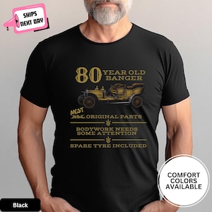 May include: Black t-shirt with a vintage car graphic and text that reads "80 Year Old Banger". The design includes the phrases "Most All Original Parts", "Bodywork Needs Some Attention", and "Spare Tyre Included".