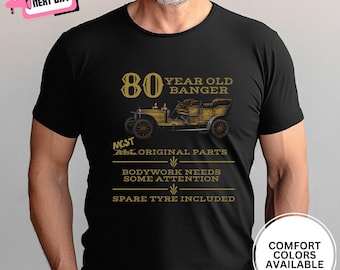 May include: Black t-shirt with a vintage car graphic and text that reads "80 Year Old Banger". The design includes the phrases "Most All Original Parts", "Bodywork Needs Some Attention", and "Spare Tyre Included".