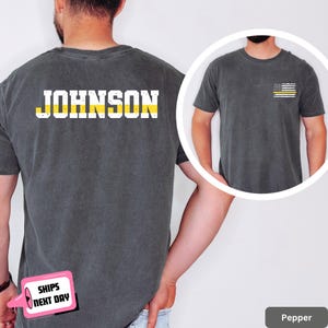 May include: Pepper-colored t-shirt with the name "JOHNSON" printed across the back in white block letters with a yellow stripe. A small American flag with a yellow stripe is on the front. The shirt is a crew neck.