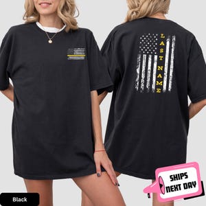 May include: Black t-shirt with a thin yellow stripe American flag on the left chest and a larger American flag with the text "LAST NAME" on the back.