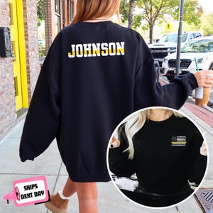 May include: Black sweatshirt with the name "JOHNSON" in white and yellow block letters across the upper back. A smaller image shows a black sweatshirt with a small American flag design with a yellow stripe on the upper left chest.