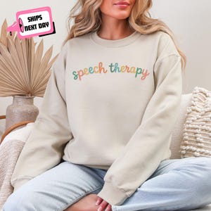May include: A beige sweatshirt with the words "speech therapy" in a colorful, rainbow-like font. The sweatshirt is a crew neck style and appears to be made of a soft material. The text is in a cursive font.