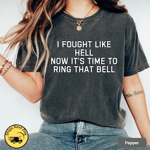 May include: A dark gray t-shirt with the text "I fought like hell now it's time to ring that bell" printed in white.