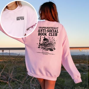 A woman wearing a pink sweatshirt with text and graphics on the back, standing in front of a scenic outdoor landscape.