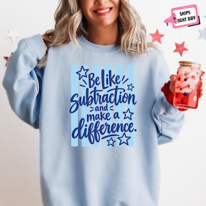 May include: A light blue crewneck sweatshirt featuring a dark blue graphic print that reads Be like subtraction and make a difference. The design includes vertical light blue stripes and several star outlines. The sweatshirt has long sleeves.