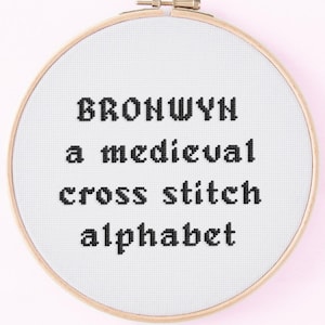 Könnte beinhalten: Ein Kreuzstich-Stickrahmen mit dem Text "BRONWYN a medieval cross stitch alphabet" in Schwarz. Die Stickerei befindet sich auf weißem Stoff, der in einem Holzrahmen eingefasst ist. Der Hintergrund ist ein zartes Rosa.