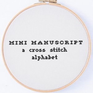 May include: A cross-stitch alphabet sampler in a wooden embroidery hoop. The text "MINI MANUSCRIPT a cross stitch alphabet" is stitched in black thread on white fabric. The hoop is round and has a brass-colored tightening mechanism.