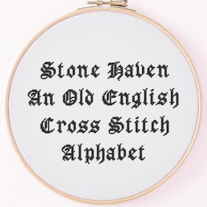 May include: A cross-stitch alphabet sampler in a wooden embroidery hoop. The text "Stone Haven An Old English Cross Stitch Alphabet" is stitched in black thread on white fabric. The hoop is secured with a brass clasp.
