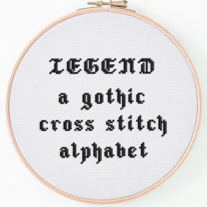 May include: A round, wooden embroidery hoop holds a white fabric with black cross-stitched text. The text reads "LEGEND a gothic cross stitch alphabet". The design is centered, with the text in a gothic font.