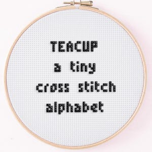 May include: A cross-stitch embroidery hoop with the words "TEACUP a tiny cross stitch alphabet" stitched in black thread on white fabric. The wooden hoop is round and secured with a metal clasp.