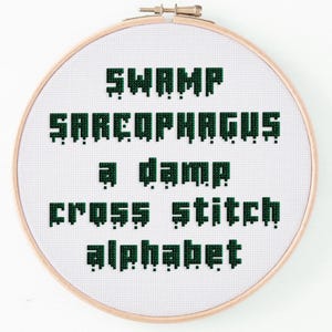 May include: A cross-stitch embroidery hoop with the words "SWAMP SARCOPHAGUS a damp cross stitch alphabet" in green pixelated font. The text is centered on a white fabric background, framed by a wooden hoop.