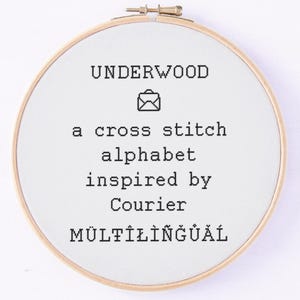 May include: A round, wooden embroidery hoop with white fabric featuring black text that reads "UNDERWOOD" above an envelope icon. Below, the text continues, "a cross stitch alphabet inspired by Courier MULTILINGUAL."