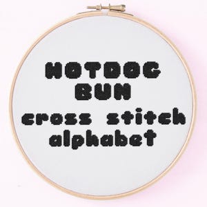 May include: A cross-stitch embroidery hoop with the words "HOTDOG BUN cross stitch alphabet" stitched in black thread on white fabric. The wooden hoop is round and has a metal clasp.