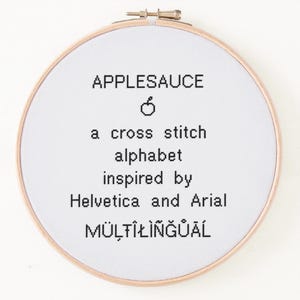 Peut inclure: Un cercle à broder avec l'inscription "APPLESAUCE", suivie de "a cross stitch alphabet inspired by Helvetica and Arial" et du mot "MULTILINGUAL". Le cercle est en bois et contient une broderie blanche.