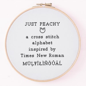 May include: A cross-stitch embroidery hoop with the words "JUST PEACHY" and "MÜLTÎLÎNGÜÂL" in black thread. The design includes a peach graphic and text that reads "a cross stitch alphabet inspired by Times New Roman". The hoop is made of wood.
