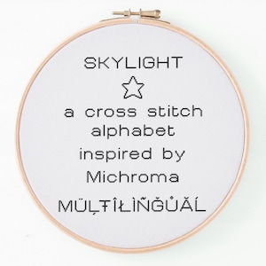 May include: A round, wooden embroidery hoop with white fabric featuring black text that reads "SKYLIGHT" above a star graphic, followed by "a cross stitch alphabet inspired by Michroma MULTILINGUAL."