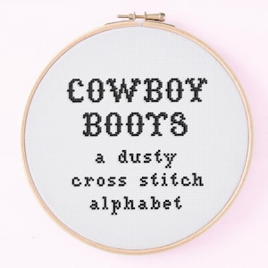 May include: A cross-stitch embroidery hoop with the words "COWBOY BOOTS" above "a dusty cross stitch alphabet" in black thread. The embroidery is on white fabric, set in a wooden hoop against a pink background.