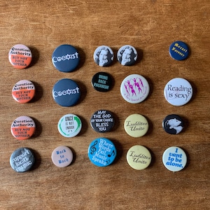 May include: A collection of 17 round button pins with various designs and slogans. The pins feature a variety of colors, including red, blue, green, yellow, and black. Some of the slogans on the pins include "Question Authority", "Coexist", "Bring Back Paganism", "Reading is Sexy", "Luddites Unite", and "Where Words Fail, Music Speaks".