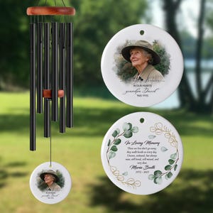 May include: A black wind chime with wooden features and three white ceramic ornaments. Two ornaments display a photo of a person with text: "In Our Hearts grandpa David 1945-2025". The other has text: "In Loving Memory Maria Booth 1972-2025".