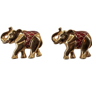 Op de afbeelding: Een paar goudkleurige olifant-oorbellen met een rood geëmailleerd zadel met een goudkleurig ruitpatroon en een kleine heldere strass-steen in het oog van elke olifant. De olifanten zijn in profiel afgebeeld, naar links kijkend, met slurf omhoog.