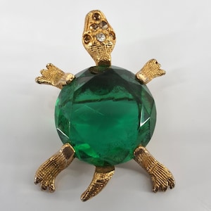 May include: A gold-toned turtle brooch featuring a large, round, faceted emerald green glass stone as the shell. The head, four legs, and tail are textured gold metal, with the head accented by small, clear, and amber-colored inset rhinestones.