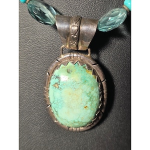 May include: A silver pendant necklace featuring a large, oval turquoise stone. The turquoise is set in a silver frame with a decorative edge. The necklace includes blue-green glass beads. The pendant hangs from a silver bail.
