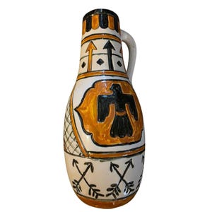 Peut inclure: Pichet en céramique avec une anse, présentant une base blanche avec des motifs géométriques orange et noirs. Le pichet comporte un emblème d'oiseau noir et des motifs de flèches. Les bords supérieur et inférieur sont décorés de bandes noires et orange.