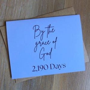 May include: A light blue rectangular greeting card with black text that reads By the grace of God 2,190 Days. The card is positioned on top of a brown kraft paper envelope. The text is written in a combination of script and serif fonts.