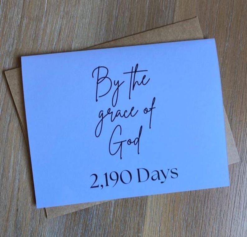 May include: A light blue rectangular greeting card with black text that reads By the grace of God 2,190 Days. The card is positioned on top of a brown kraft paper envelope. The text is written in a combination of script and serif fonts.
