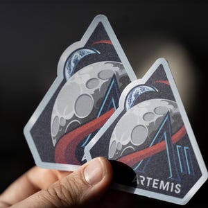 Könnte beinhalten: Zwei Artemis-Aufkleber werden gehalten. Jeder Aufkleber zeigt einen stilisierten Mond mit Kratern, eine Mondsichel-Erde und rote und blaue Linien. Das Wort "ARTEMIS" ist blau gedruckt.