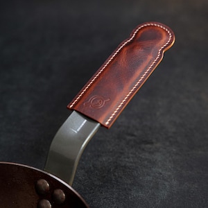 May include: Close-up of a dark brown leather handle cover stitched with white thread, attached to a metal pan handle. The leather has a logo embossed on it. The pan is dark brown.