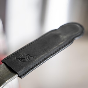 May include: Close-up of a black leather handle cover with a stitched edge and a debossed circular logo. The cover is attached to a metal object, possibly a kitchen utensil or tool.