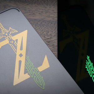 Puede incluir: Un portátil gris con un logotipo de Zelda dorado, con una espada y la letra "Z". Una segunda imagen muestra el logotipo brillando en verde en la oscuridad. El diseño es una representación estilizada de un videojuego.