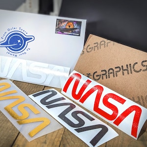 May include: An assortment of NASA-themed stickers in gold, black, and red, alongside a white envelope with a blue and white space-themed logo and the text "From Outer Space To Your Home!" A brown cardboard box with the word "GRAPHICS" is also visible.