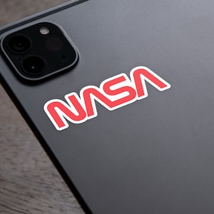 Puede incluir: Una pegatina con el logotipo de la NASA en rojo con borde blanco está adherida a una tableta gris oscuro. El texto de la NASA está en negrita y sin serifa. La tableta tiene una protuberancia de la cámara en la esquina superior izquierda.