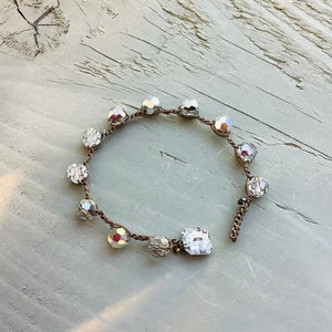 May include: A silver and brown beaded bracelet with a square-shaped charm. The bracelet is made with a brown cord and features round silver beads.