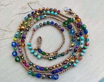Puede incluir: Collar de múltiples hilos con una variedad de cuentas de colores, incluyendo turquesa, azul, verde y dorado. El collar está hecho de cordón marrón y tiene un dije de cristal en el extremo.