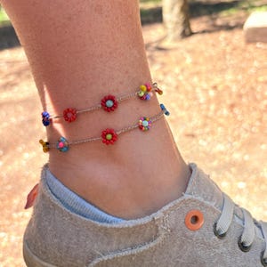 May include: Two delicate anklets made of thin beige cord featuring small, colorful beaded flower motifs. Each flower is constructed from tiny round beads in shades of red, blue, yellow, and pink, spaced evenly along the cord to create a floral pattern.