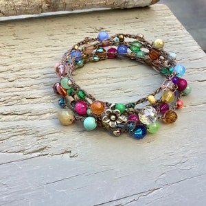 May include: Multicolored beaded bracelet with a silver flower charm. The bracelet is made of crocheted thread and features a variety of beads, including turquoise, pink, green, and blue.