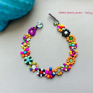 Könnte beinhalten: Ein farbenfrohes Perlenarmband mit Gänseblümchen-Blütenmustern in verschiedenen Farben, darunter Orange, Lila, Grün und Blau. Das Armband hat einen transparenten Verschluss und den Text "Pebble Beach Jewels - Daisy bracelet".